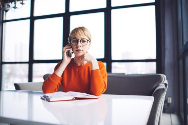 Ciddi bir uzaktan kumandalı kadın çalışanın resmi. Masada oturmuş, açık defterle kameraya bakıyor ve telefon görüşmesi yaparken yüzüne dokunuyor.