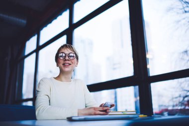 Düşük açılı, dişlek gülümsemeli bir kadın masada tek başına otururken masaya oturmuş kameraya bakıyor.