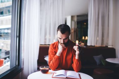 Konsantre sakallı erkek elinde bir fincan sıcak çayla oturmuş kitap okurken ve elinde gözlük tutarak aydınlık odada boş vakit geçirirken aşağıya bakıyor.