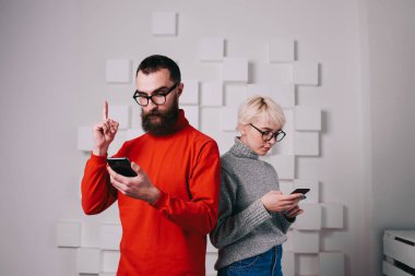 Odaklanmış sakallı, turuncu kazaklı, gözlüklü, akıllı telefon kullanan ve akıllı telefondan mesaj okuyan sarışın kadını işaret eden genç bir adam.