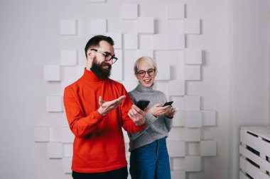 Neşeli genç sakallı adam cep telefonuyla komik videolar gösteriyor. Beyaz duvarlı, geometrik panelli, gülümseyen bir sarışına.