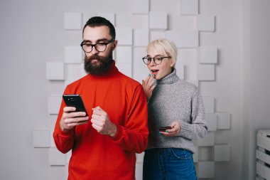 Gri süveterli ve gözlüklü genç bir bayan genç hippinin omzundan akıllı telefondan video izliyor ve şaşırmış görünüyor.