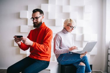 Neşeli adam cep telefonundan video izliyor boş zamanı var ve ciddi bir kadın ışık odasında çalışırken dizüstü bilgisayarda yazıyor.