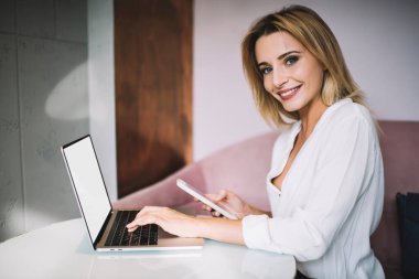 Beyaz tişörtlü pozitif bir genç bayanın yan görünüşü odadaki kanepenin yanındaki masada bilgisayarın başında çalışıyor ve cep telefonundan bildirimleri kontrol ediyor ve kameraya bakıyor.