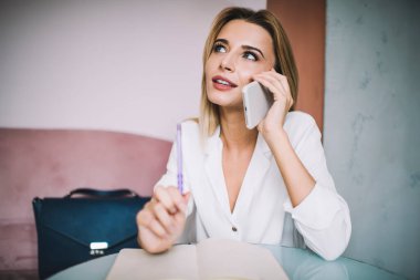 Beyaz bluzlu düşünceli genç bir bayan akıllı telefonla konuşurken başını kaldırıp evdeki yuvarlak masada oturup not alıyor.