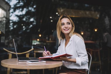 Gündüz vakti kafenin terasında dizüstü bilgisayar ararken not defterine not alıp başka yere bakan neşeli bir girişimci kadın.