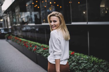 Beyaz bluzlu, gülümseyen sarışının arka görüntüsü. Sokakta modern bina boyunca yürüyor ve akşamları omzunun üzerinden bakıyor.