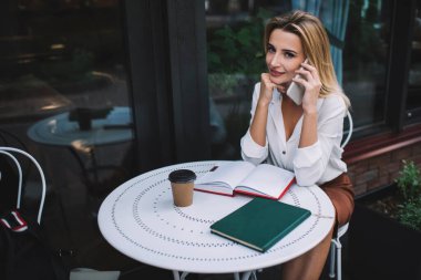 Yuvarlak masada oturan zarif giysili karizmatik bayandan açık defter ve kahve alıp kafenin terasında akıllı telefondan konuşan.