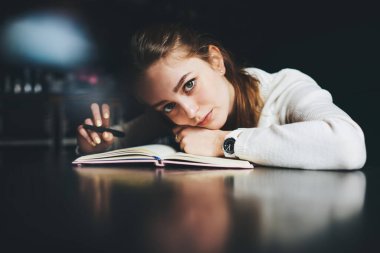 Beyaz kazaklı, genç ve ciddi bir kadın serbest çalışan planlamacıyla masaya yaslanıp ertesi gün için plan yapan kameraya bakıyor.