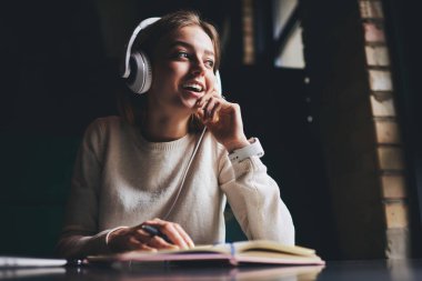 Gündelik kıyafetlerle ve kulaklıklarla gezen mutlu bir kadın zamanını geçirirken müzik dinliyor ve modern çalışma alanındaki not defterine not alıyor.