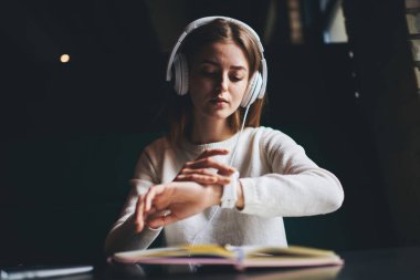 Günlük kıyafetleri ve beyaz kulaklıkları olan konsantre bir kadın kitap okurken kol saatini kontrol ediyor ve alacakaranlıkta yaratıcı odada müzik dinliyor.