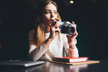 Günlük giysiler içindeki konsantre kadın fotoğrafçı gözlerini kaçırıyor ve kapalı dizüstü bilgisayarı ve defteri olan siyah masada otururken ağzı açık eski fotoğraf makinesiyle fotoğraf çekiyor.
