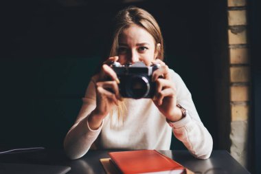 Genç, gülümseyen kadın fotoğrafçı. Analog fotoğraf makinesiyle fotoğraf çekiyor. Masada kitap ile birlikte oturuyor ve kameraya bakıyor.