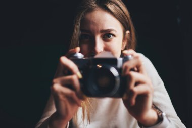 Beyaz kazaklı, genç, neşeli kadın fotoğrafçı. Retro kamerada fotoğraf çekmeye hazırlanıyor ve karanlık odada kameraya bakıyor.