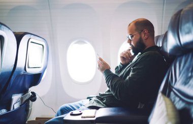 Gözlüklü ve günlük kıyafetli ciddi bir yetişkinin uçakta pencerenin yanında oturması ve telefondaki uyarıları kontrol etmesi.