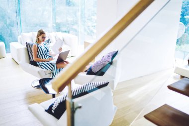 Genç serbest çalışan bir bayanın elinde belgelerle koltukta oturması ve açık oturma odasındaki dizüstü bilgisayarında yeni bir iş projesi üzerinde çalışması.