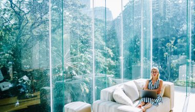 Kırmızı dudaklı, kameraya bakan ve bilgisayar kullanan mutlu kadın serbest yazar. Çağdaş çalışma alanında cam duvarlı rahat bir koltukta dinleniyor.