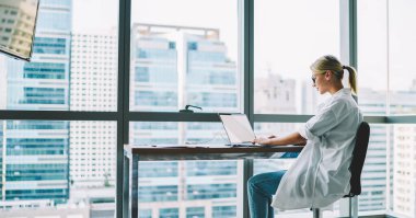Beyaz tişörtlü ve gözlüklü bir kadının modern ofiste dizüstü bilgisayarla çalışırken gündüz duvar manzarasına karşı kenar görüntüsü