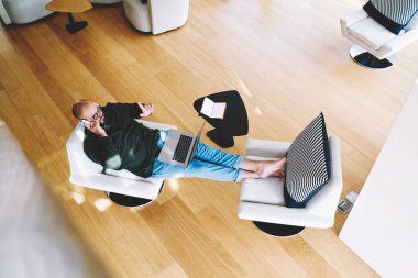 Dizüstü bilgisayarla koltukta oturan, akıllı telefondan konuşan ve geniş bir odada el kol hareketleriyle hareket eden serbest erkek çalışanlardan.