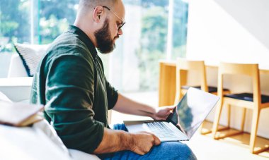 Güneşli bir günde cam terasta dizüstü bilgisayarda video izleyen günlük giysili ve gözlüklü genç adamın yan görüntüsü