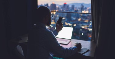 Tişörtlü, dizüstü bilgisayarlı ve akıllı telefonlu bir erkeğin modern ofiste not defterine not aldığı arka plan görüntüsü.