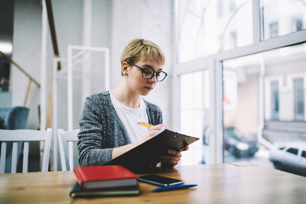 Concentrated female worker in casual clothes with eyeglasses sitting at table near window using clipboard and writing notes working in modern office