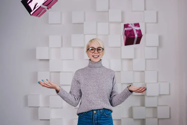 笑顔ブロンド女性でカジュアルセーターと眼鏡見カメラと実証浮動プレゼントボックスでホワイト現代的な部屋 — ストック写真