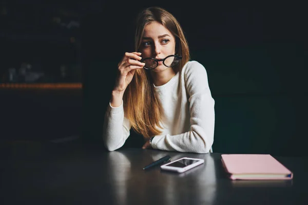 若いです深刻な女性でカジュアル服を保持眼鏡離れて携帯電話やノートブックとテーブルに座って夜 — ストック写真