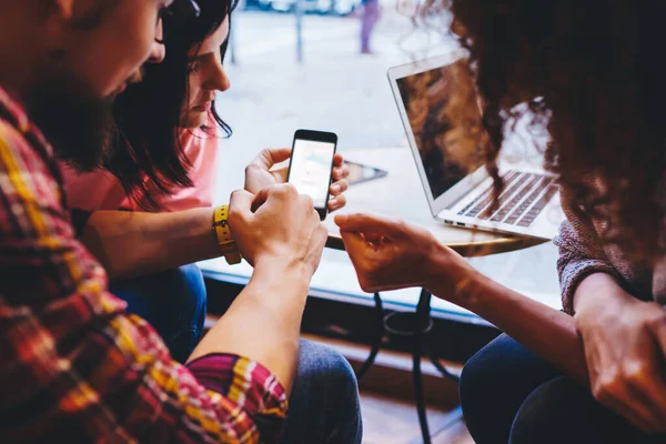 Crop unrecognizable guy with mobile phone browsing and showing photos to female friends while sitting together near table with laptop in daytime