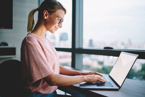 Side view of happy executive female in casual outfit and eyewear sitting near window and typing on laptop while working on project at sundown