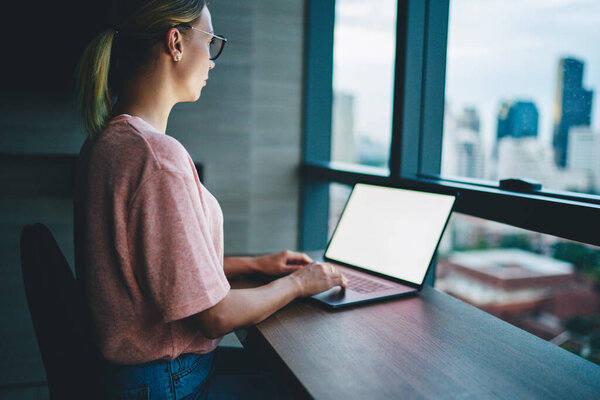 Side view of crop female entrepreneur in casual wear and eyeglasses typing on laptop while working on business strategy in modern office at sunset