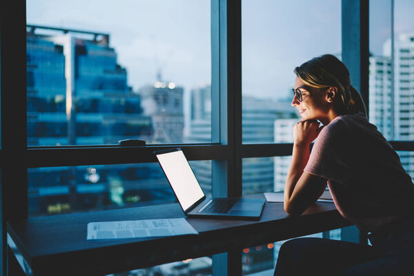 Side view of female worker with toothy smile sitting at table with laptop looking at screen in modern office in twilights