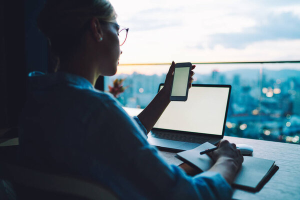 Back view of crop anonymous female in formal wear and eyeglasses browsing smartphone and taking notes while working on laptop in modern office at sunset