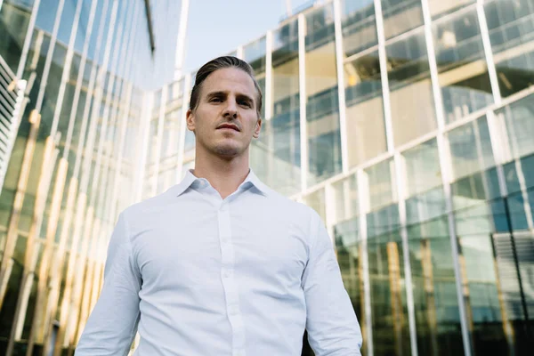 From below view of confident male entrepreneur in formal shirt standing outside modern office building and looking at camera pensively