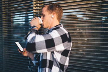 Gündelik giyinen, kulaklıkla müzik dinleyen ve dışarıda kahve içerken akıllı telefon kullanan genç bir erkeğin yan görüntüsü.