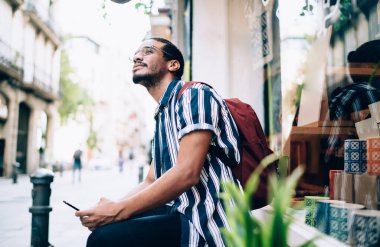 Düşünceli, sakallı, sıradan giyinmiş, akıllı telefon kullanan ve gündüz vakti kafasını kaldırıp bakan etnik bir adamın yan görüntüsü.