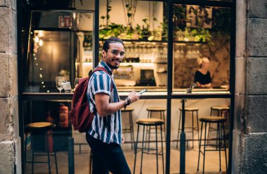 20 'li yaşlarda genç, koyu tenli bir erkek. Şehirde gezinmek için navigasyon uygulaması kullanıyor. Şehir caddesinde yürüyor.