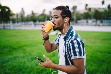 Modayı takip eden, modaya uygun saç kesimi olan ciddi etnik erkeklerin yan görünüşü. Parkta yürürken kahve içip, cep telefonlarını karıştırmak.