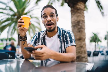 Gözlüklü heyecanlı hippi adam kameraya bakıyor. 4G internete bağlı cep telefonu tutuyor. Afro-amerikan kahvesi içiyor ve boş zamanlarında akıllı telefon kullanıyor.