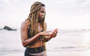 Koyu tenli bayan sporcu, sabah için mobil uygulamadan müzik seçiyor. Açık havada koşuyor, kulaklıkla kulaklıkla akıllı telefondan müzik dinliyor.