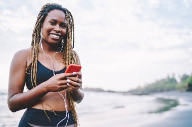 Müzik podcast 'i seçerken kameraya gülümseyen modern hücresel cihazlı neşeli fit bir kızın yarım boy portresi. Elinde akıllı telefon cihazı olan ve poz veren koyu tenli mutlu koşucu.