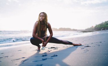 Sahil şeridinde sabah pilates antrenmanı sırasında bacak kaslarını esneten formda formda bir kızın portresi. Plaj eğitiminde eşofman giyen esnek bir bayan sporcu ısınıyor.