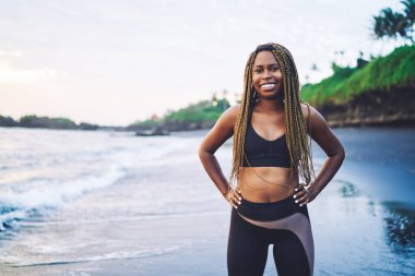 Aktif giyinmiş neşeli Afro-Amerikan kadın sabah vakti deniz kıyısında gülerek, kamera karşısında gülümseyen koyu tenli kadın koşucunun yarım boy portresi.