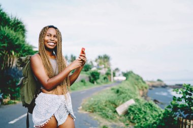 Sırt çantalı neşeli Afrikalı Amerikalı gezgin akıllı telefon uygulaması ve internet kullanarak tropikal çevre fotoğrafları çekiyor, yaz gezisi sırasında neşeli turist manzarası çekiyor.