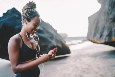 Mobil teknolojisi olan neşeli formda bir kız. Sahil şeridinde sabah antrenmanı yaparken medya içeriğini kontrol ediyor. Elektronik kulaklıklı mutlu bir bayan sporcu sosyal ağlarda akıllı telefon mesajlarıyla eğleniyor.