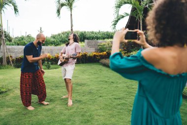 Birlikte gitar çalarken gülümseyen ve eğlenen mutlu çok ırklı arkadaşlarla dolu bir vücut ve onların fotoğraflarını çeken bir kadın.