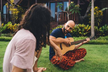 Yoğunlaşmış sakallı, gündelik giyimli, güneşli bir günde bahçedeki yeşil çimlerde oturan ve arkadaşıyla vakit geçirirken gitar çalan etnik bir adam.