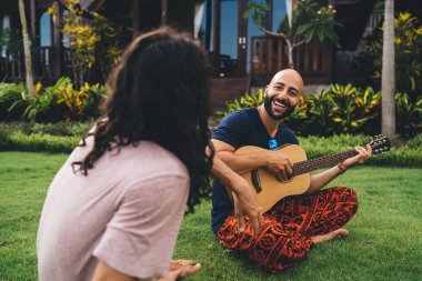 Neşeli, sakallı, sıradan kıyafetli, yeşil çimlerde oturan ve arkadaşlarıyla bahçede takılırken gitarın tadını çıkaran bir etnik adam.