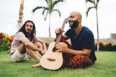 Çimenlerin üzerinde oturan genç adam tropikal yeşil çayırlarda gitar çalıyor. Avuçlar birbirine sarkıyor ve başka tarafa bakıyor.
