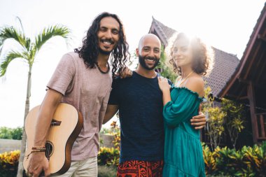 Düşük açılı pozitif çoklu etnik arkadaş grubu gündelik kıyafetleriyle gitarla gülümseyip dinlenirken güneşli bir günde sarılıp kameraya bakıyor.